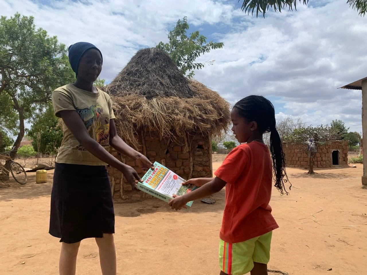 A girl handing a Books & Beyond leaflet to a younger
                    child outside a thatched home in rural Makueni