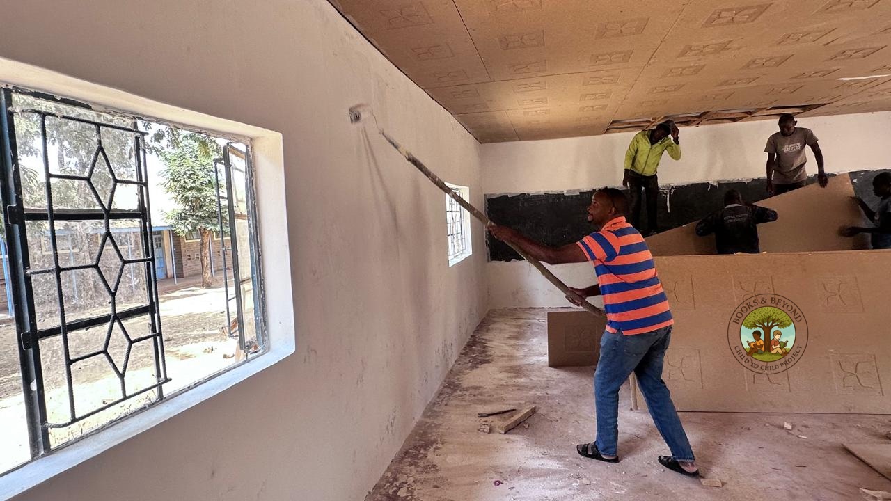 At work — rolling white paint across the library wall, ceiling boards freshly fitted above, the school grounds visible through the window. Progress, one coat at a time.